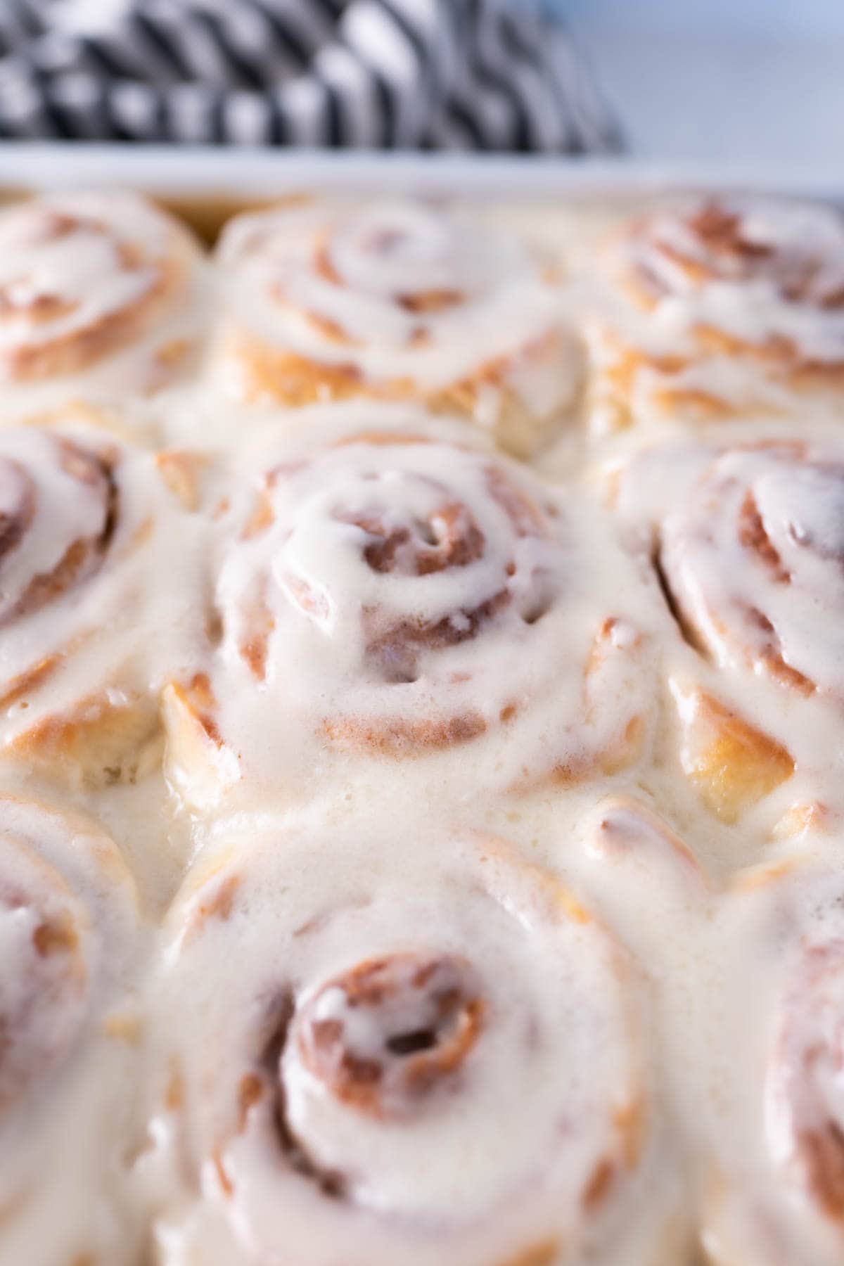 frosted or iced cinnamon rolls in a pan with a striped dishtowel behind them. 