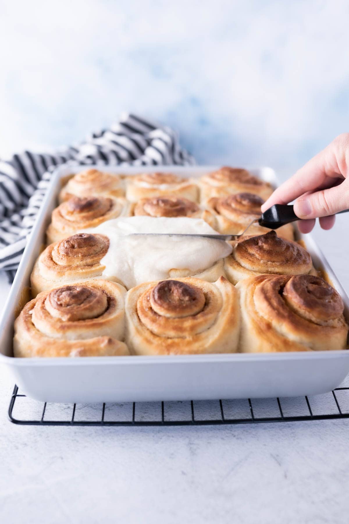 cinnamon roll icing being spread onto warm cinnamon rolls with an offset spatula. 