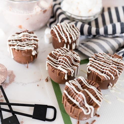 hot chocolate bombs, drizzled with white and milk chocolate on the counter with hot cocoa in the background