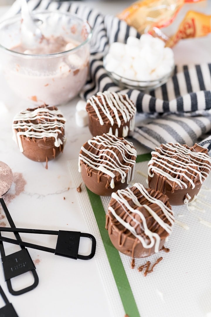 hot chocolate bombs, drizzled with white and milk chocolate on the counter with hot cocoa in the background 