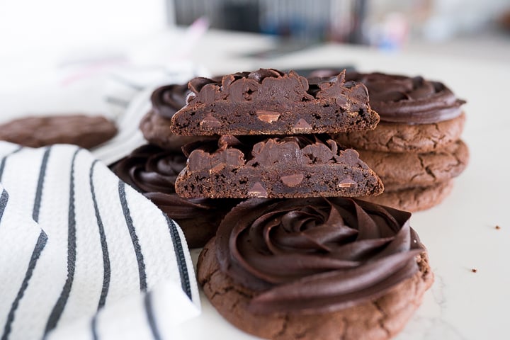 chocolate cookies with chocolate fudge frosting on top on the counter. One cookie is cut in half and stacked on top of each other to show the inside of the cookies.