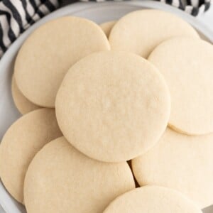 circle sugar cookies, piled onto a plate with a. striped dishtowel behind them.