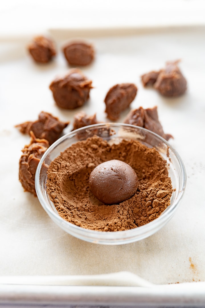 A chocolate truffle in a bowl of cocoa powder with chocolate truffles in the background on parchment paper.
