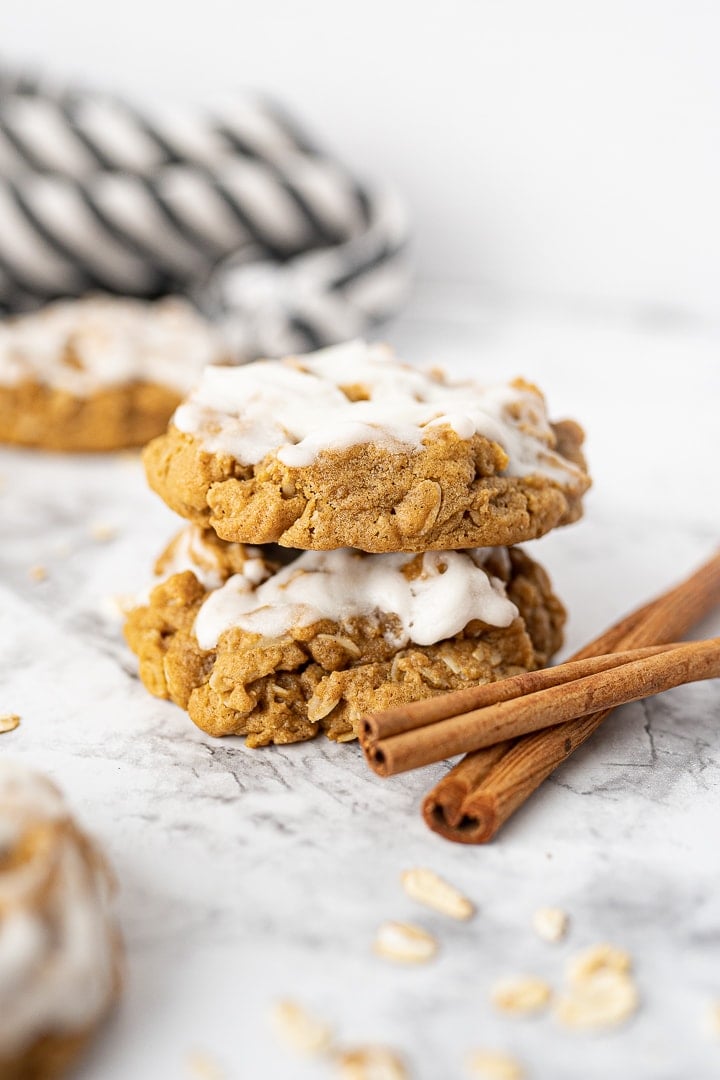 Two iced oatmeal gingersnap cookies stacked on the counter with cinnamon sticks and a black and white striped dish towel in the background.