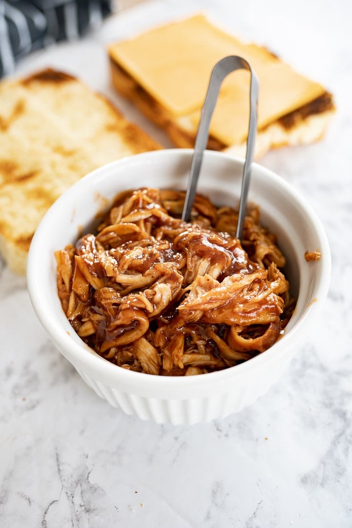 A small white bowl of pulled chicken on the counter with open face rolls, topped with cheese, in the background.