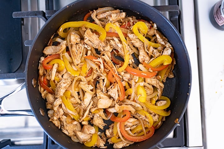 A pan on the stove filled with bell peppers, chicken, and onions.