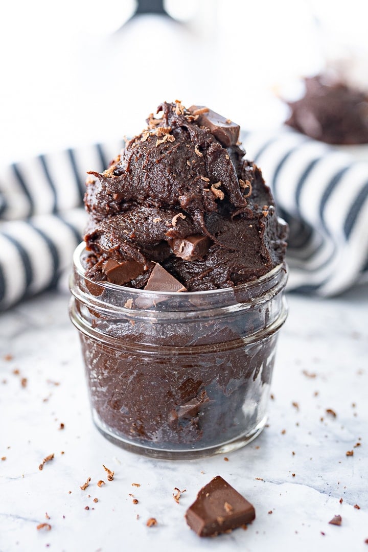 A glass jar of brownie batter stuffed with large chocolate chunks, on the counter.