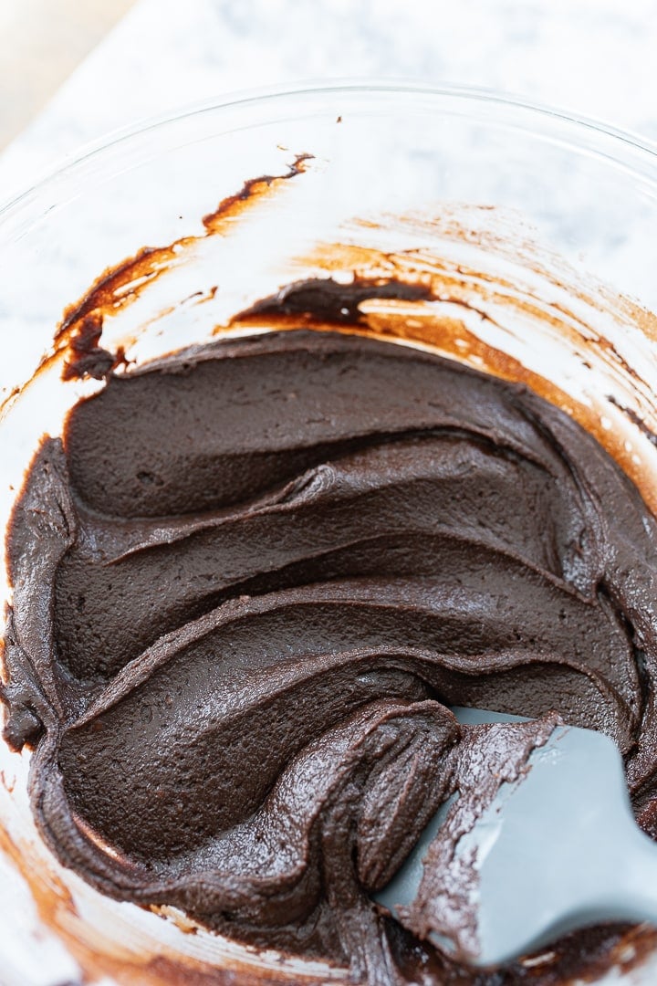 A glass bowl with creamy brownie batter and a grey spatula stirring the batter.