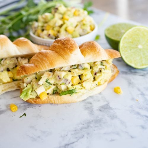 avocado chicken salad on a croissant with avocado, chicken, corn, cilantro and lime on the counter.
