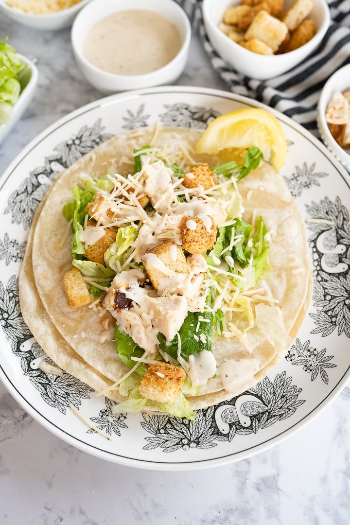 Chicken caesar filling on an open tortilla sitting on a china plate. Bowls with caesar dressing and croutons above it resting on a black and white striped towel.  