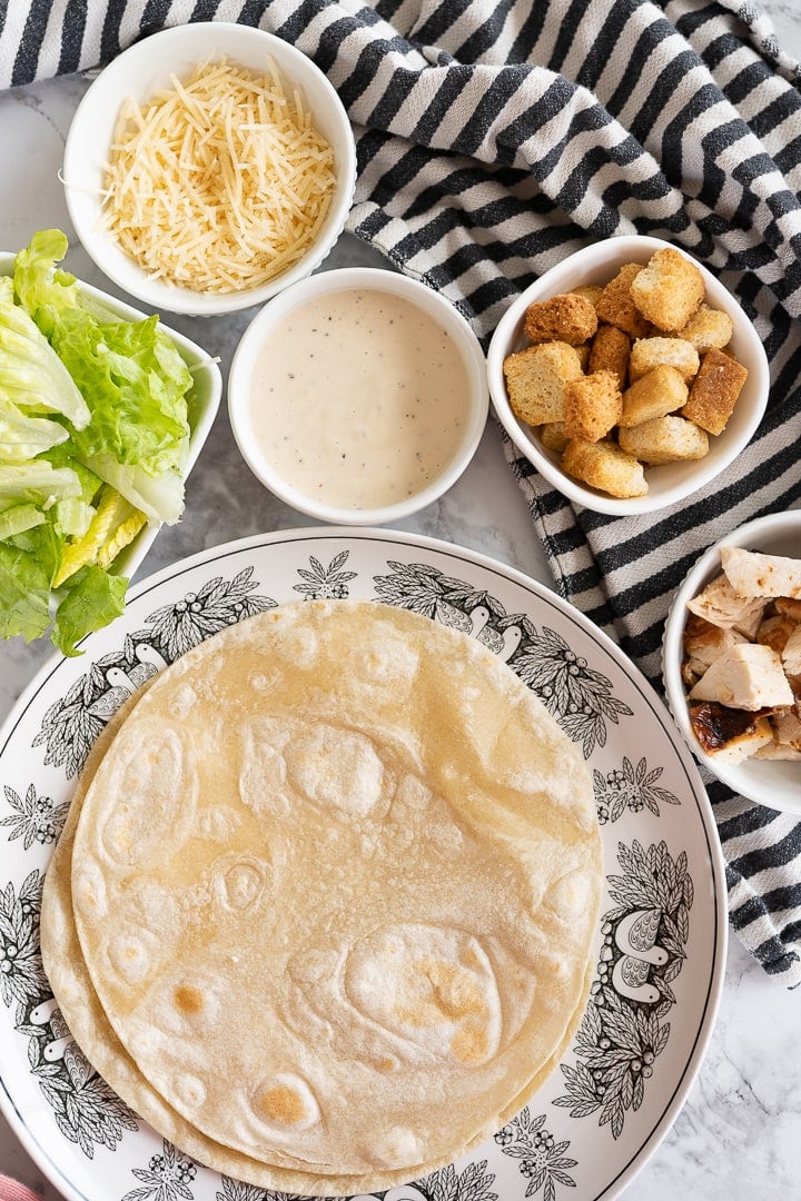 A stack of tortillas rests on a plate in the bottom left. To the upper right there is a striped black and white towel with bowls of croutons, dressing, cheese and romaine lettuce. 