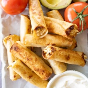 Chicken taquitos piled high in the center of the photo. At the top, there are two whole tomatoes and half of an avocado. At the bottom there is a bowl of sour cream.