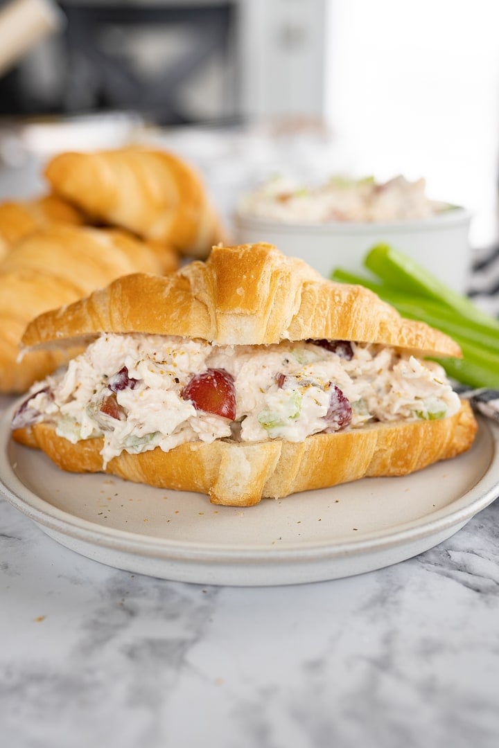 A chicken salad croissant sandwich sits on a plate. In the background you can see additional celery and croissants. 