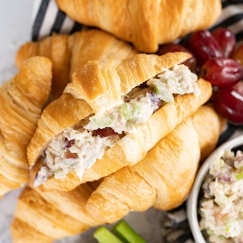 A stuffed chicken salad sandwich is on it's side with the filling facing up. Sandwich sits on additional crossiants, celery and grapes. Across the top it says "5 ingredient chicken salad sandwiches"