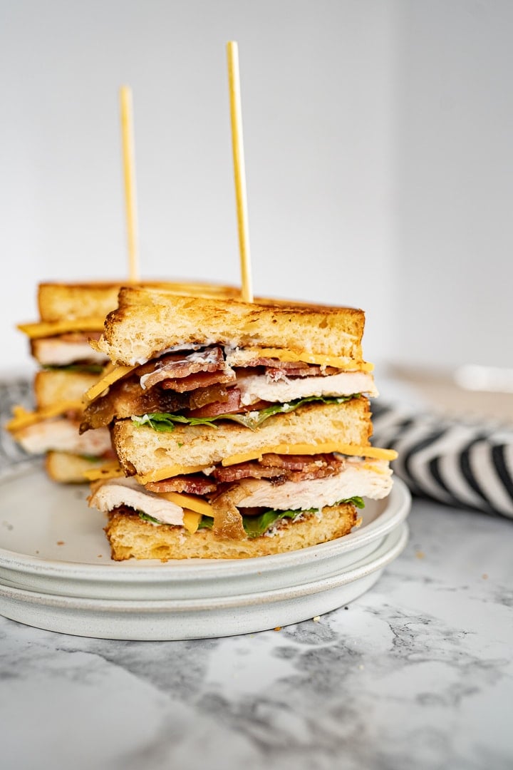 Two double decker chicken club sandwiches sit on a glass plate. They have toothpicks holding the sandwiches together. Next to the plate is a black and white striped towel. 