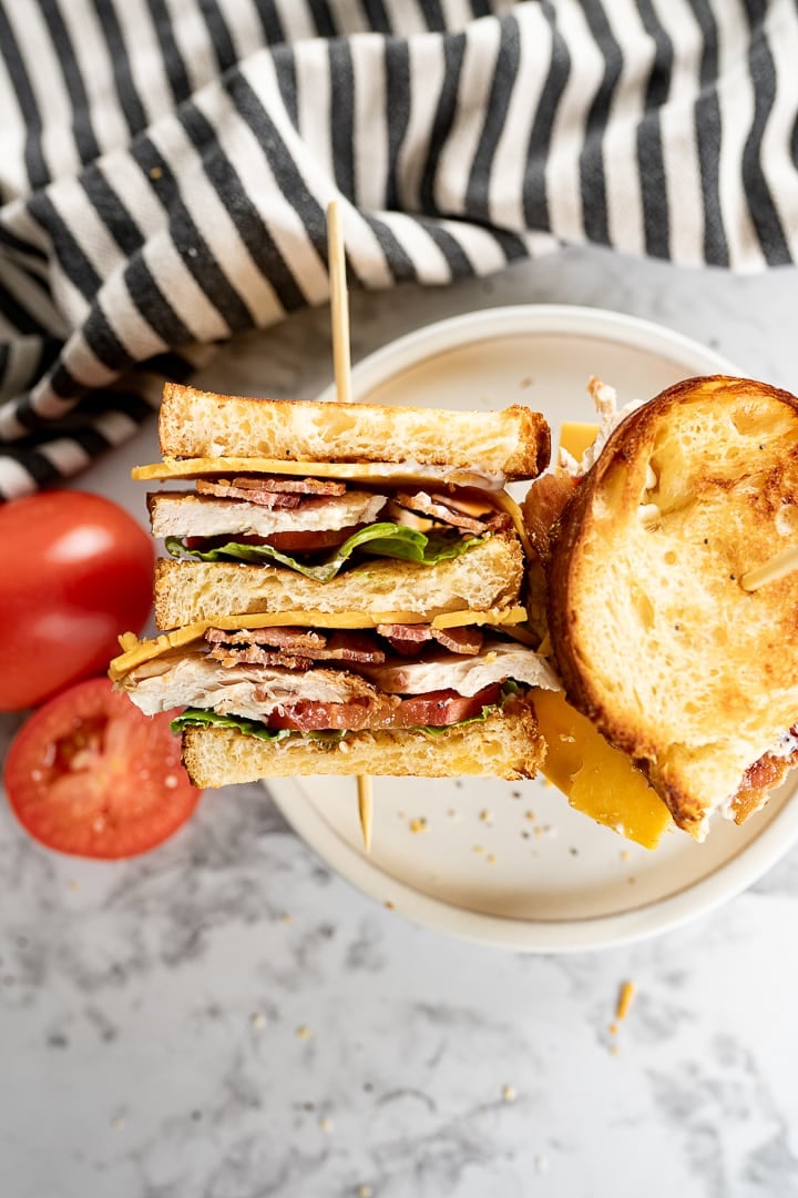 An aerial view of a plate with two halves of a chicken club sandwich on it. One half is on it's side with the fillings facing up while the other is sitting normally. Next to the plate is a whole tomato and a halved tomato and a black and white striped towel. 