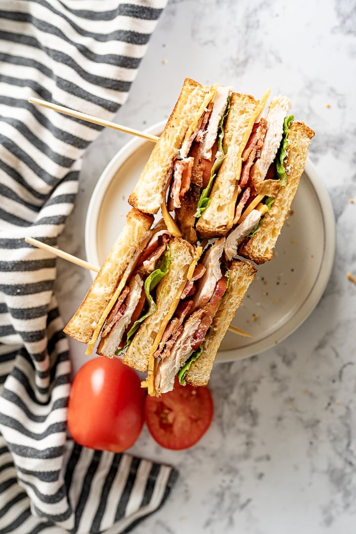 Two halves of a chicken club sandwich are laying on their side on a plate. Next to the plate is a sliced tomato and a black and white striped towel. 