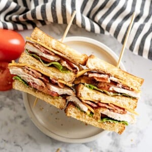 Two halves of a chicken club sandwich are laying on their side on a plate. Next to the plate is a sliced tomato and a black and white striped towel.