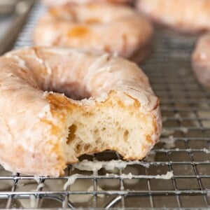 A glazed donut with a bite out of it on a wire cooling rack. In the background you can see additional donuts.