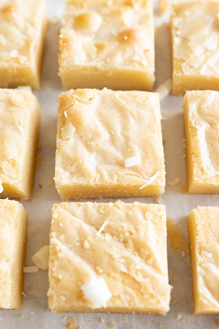 9 squares of white chocolate brownies are lined up in a grid on the counter. There are white chocolate flakes sprinkled on top. 