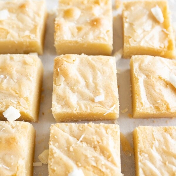 9 squares of white chocolate brownies are lined up in a grid on the counter. There are white chocolate flakes sprinkled on top.