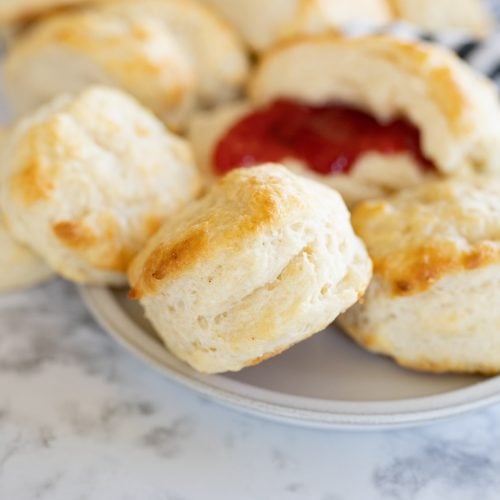 Golden buttermilk biscuits on a plate. One of the biscuits is open with jam on it.
