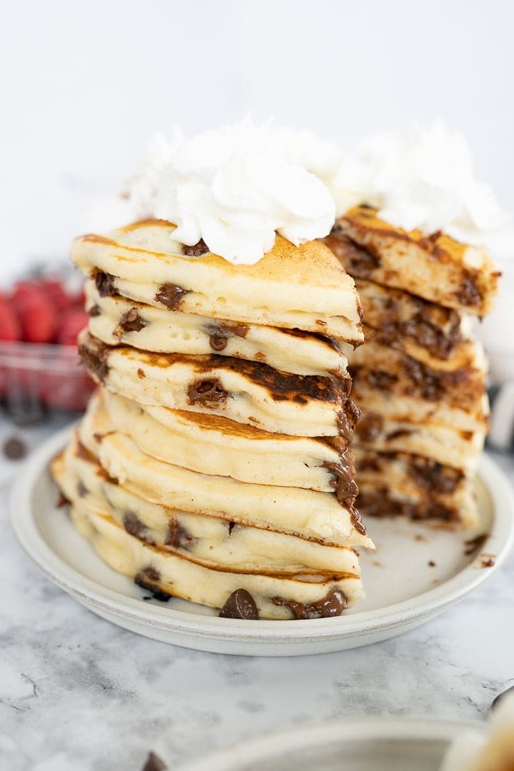 A stack of chocolate chip pancakes with a quarter cut out. Topped with whipped cream. 