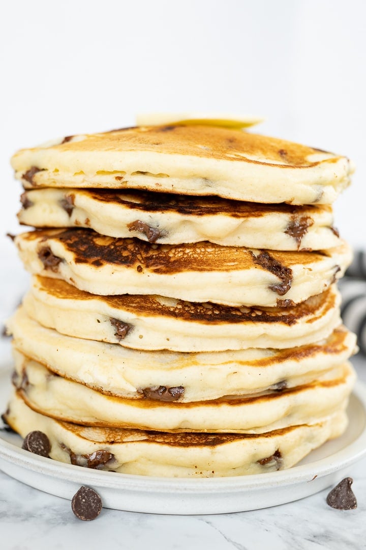 A tall stack of chocolate chip panckaes on a plate with chocolate chips scattered around. 