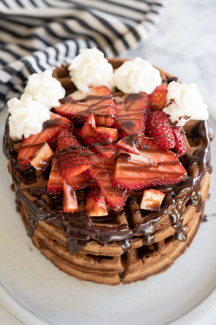 Aerial view of a stack of chocolate waffle topped with piped whipped cream, chocolate sauce and chopped strawberries. 