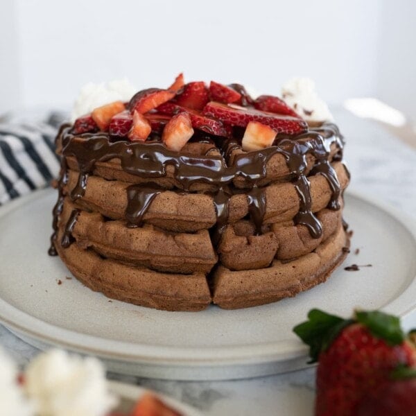 Chocolate waffles stacked high on a plate. Topped with chocolate syrup an dcut up strawberries.