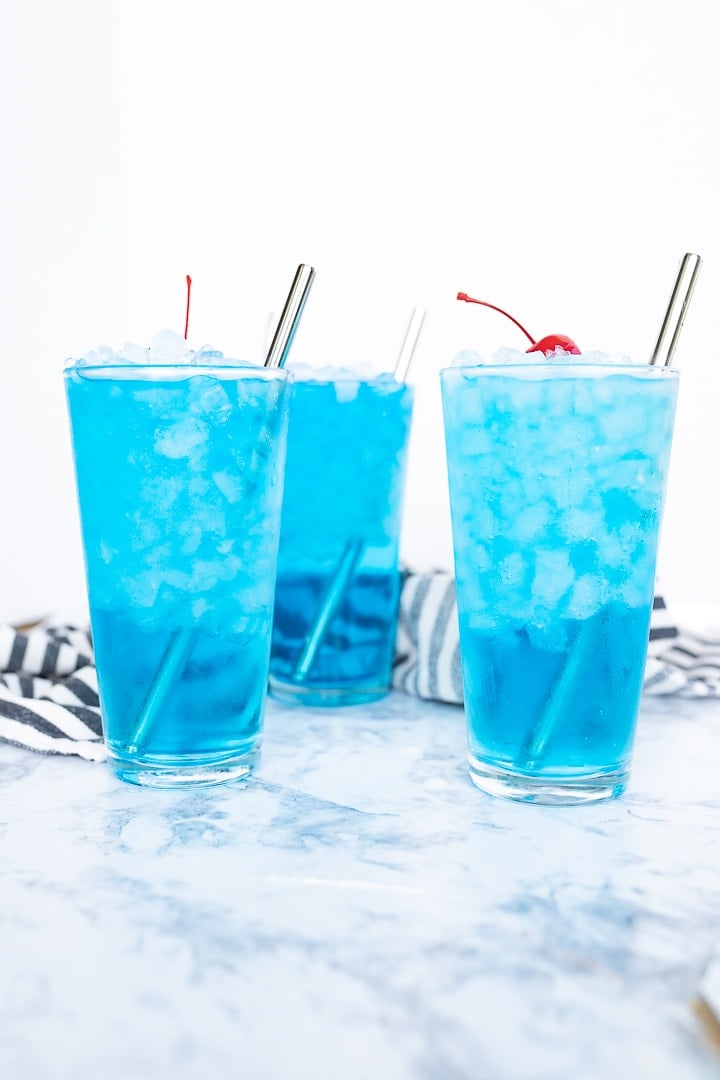 Side view of 3 glasses of blue sonic ocean water, topped with maraschino cherries and served over pebble ice. 
