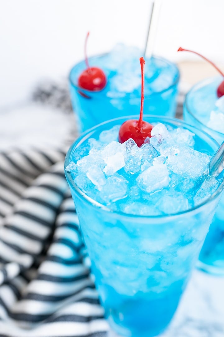 Partial view of 2 glasses of blue sonic ocean water with maraschino cherries and stainless steel straws. In the background is a black and white striped towel.