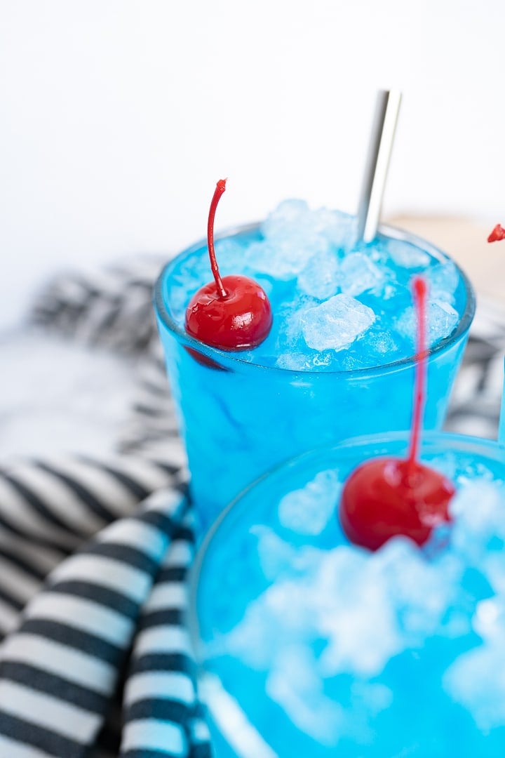 Partial view of 2 glasses of blue sonic ocean water with maraschino cherries and stainless steel straws. In the background is a black and white striped towel. 