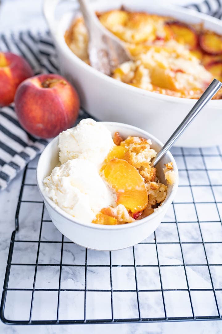 A bowl of homemade peach cobbler topped with ice cream and a spoon sits on a counter next to a bigger serving dish of the cobbler. 