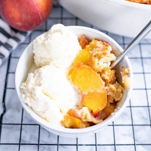 A bowl of homemade peach cobbler topped with ice cream and a spoon sticking out of it.