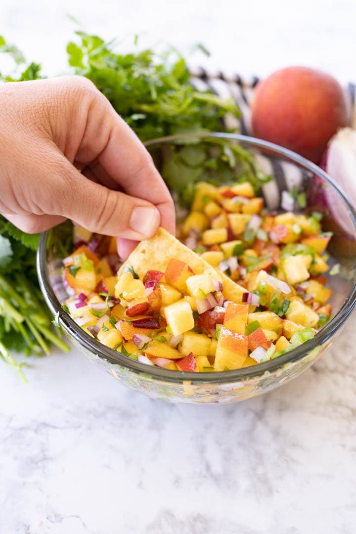 A bowl of peach salsa with a hand dipping a chip into it. 