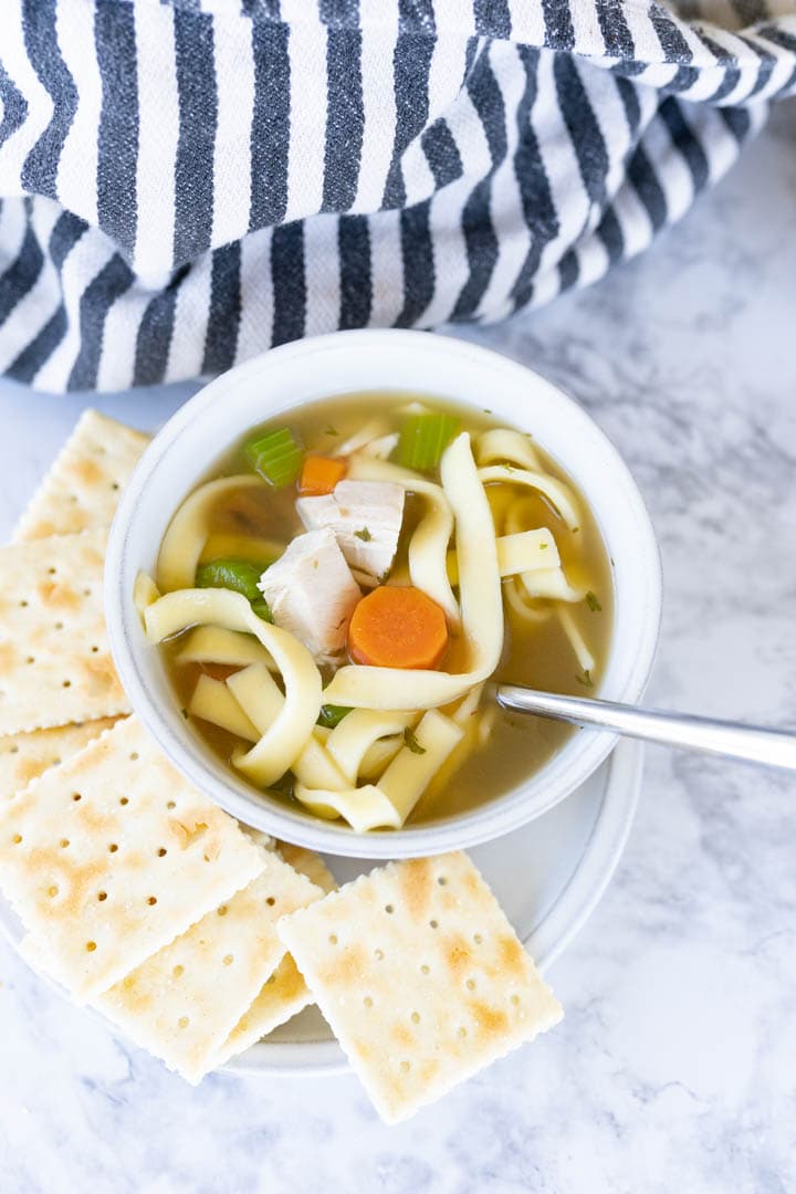 Aerial view of chicken noodle soup in a bowl with a spoon sticking out of it. Crackers are next to it. 