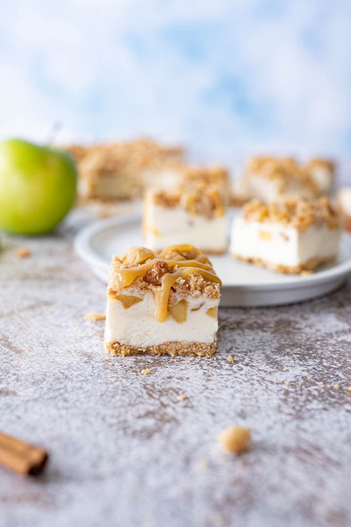 A caramel apple cheesecake bar sitting on the counter next to a plate filled with additional bars and a green apple.