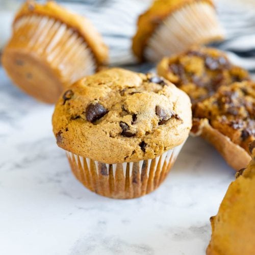 A countertop filled with pumpkin chocolate chip muffins. Some are turned on their side and others have been opened to show the gooey chocolate chips.