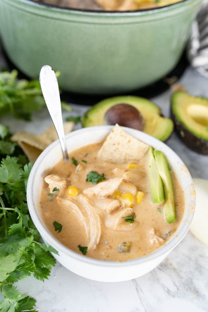 A single serving bowl of white chicken chili with a spoon sticking out of it. Next to it on the counter are cilantro and avocado garnishes. 