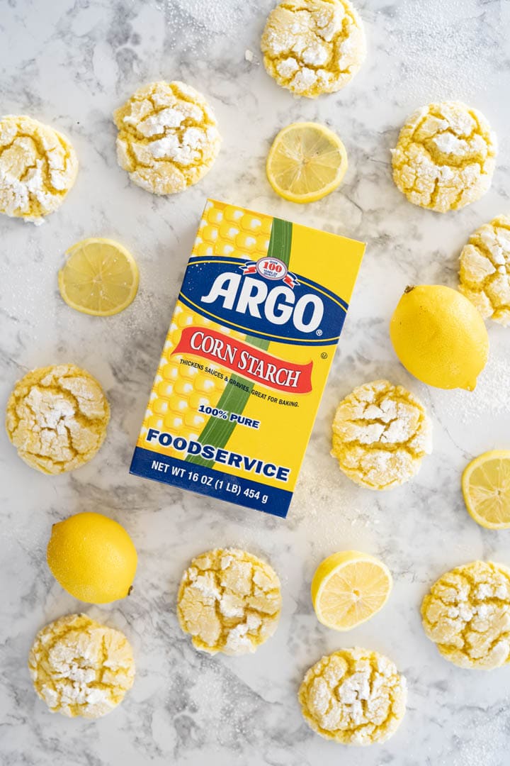 Lemon crinkle cookies along with some lemon slices on the counter surrounding a box of Argo corn starch, photo taken from an overhead angle.