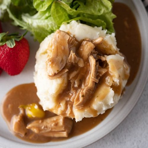 Mississippi Chicken served over mashed potatoes on a plate with salad and a strawberry.