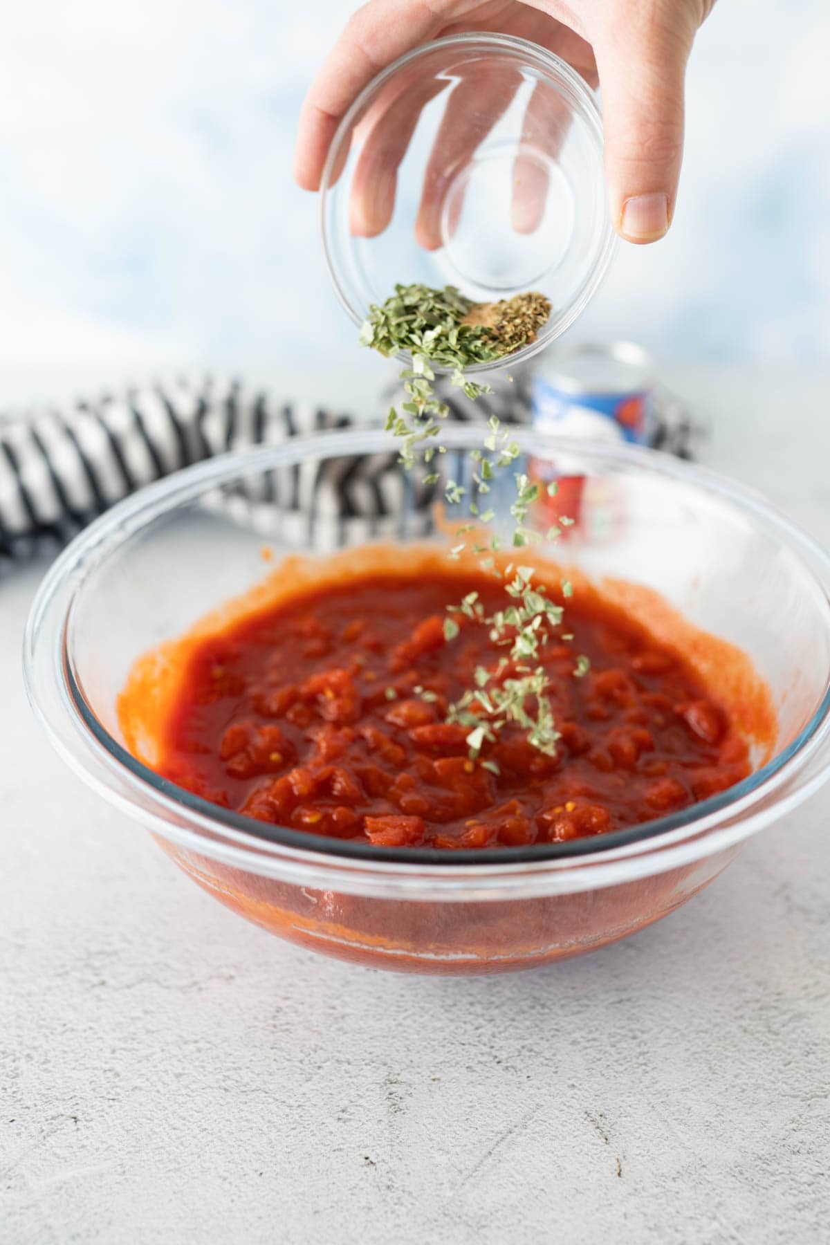A bowl of pizza sauce with seasonings being dumped in. 