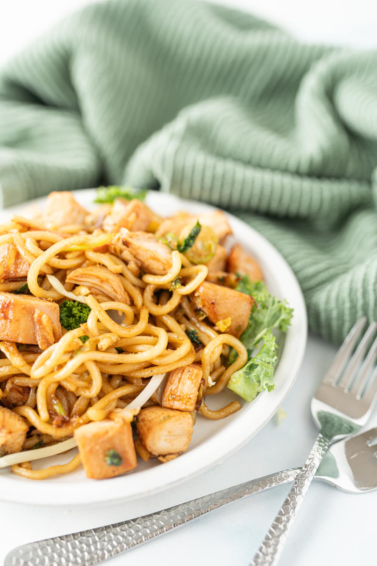 Side view of a bowl of chicken chow mein on a counter with a green towel and silverware. 
