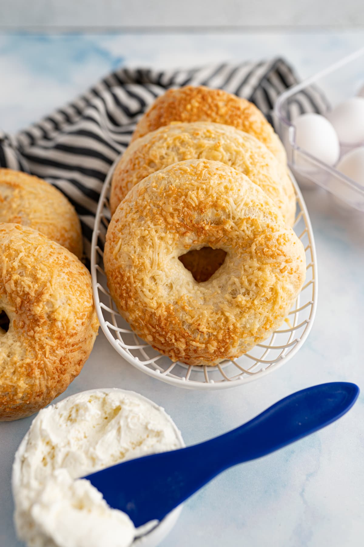 asiago cheese bagels in a wire basket next to some cream cheese and a black and white striped dishtowel. 