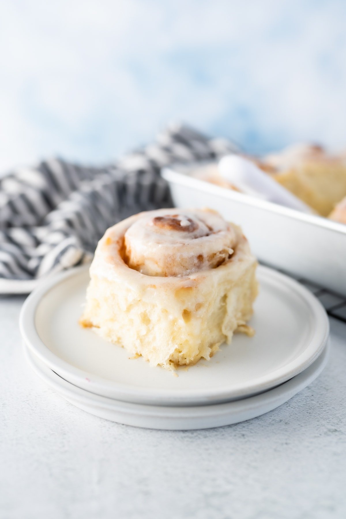 Side view of a fluffy homemade cinnamon roll on a plate next to a baking pan of extra rolls. 