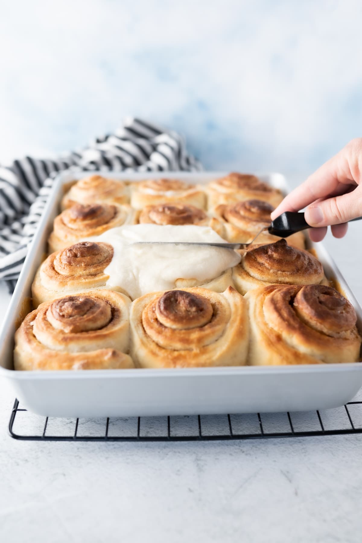 A pan of baked cinnamon rolls being frosted. 