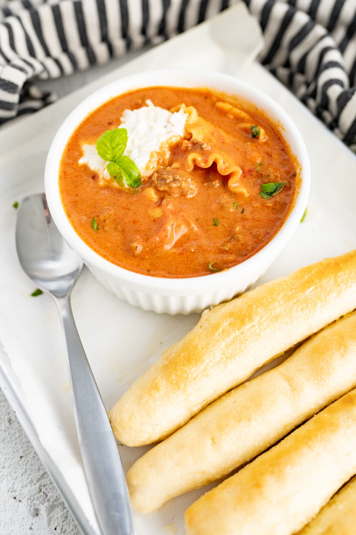 A bowl of lasagna soup next to a couple of breadsticks. 