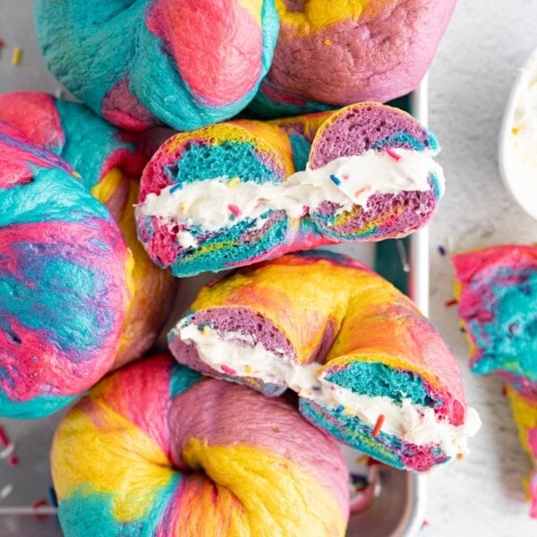 A counter filled with rainbow bagels. One bagel is cut in half with cream cheese in the middle.