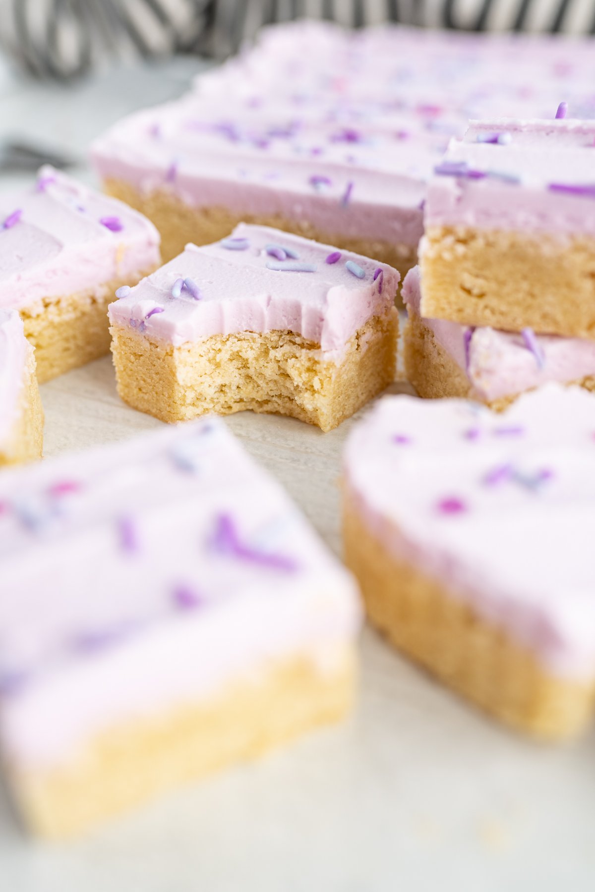 A pile of sugar cookie bars frosted with purple frosting and topped with sprinkles. One bar has a bite taken out of it. 